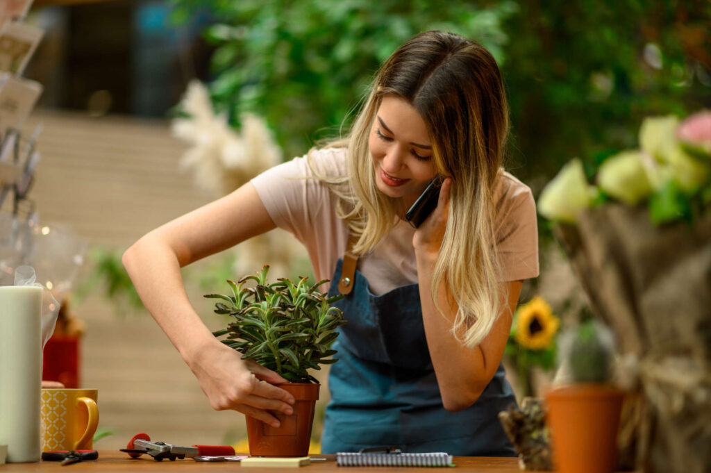 Florist talking with client on the smartphone and taking an order while standing next to the counter surrounded with flowers and plants in a flower shop